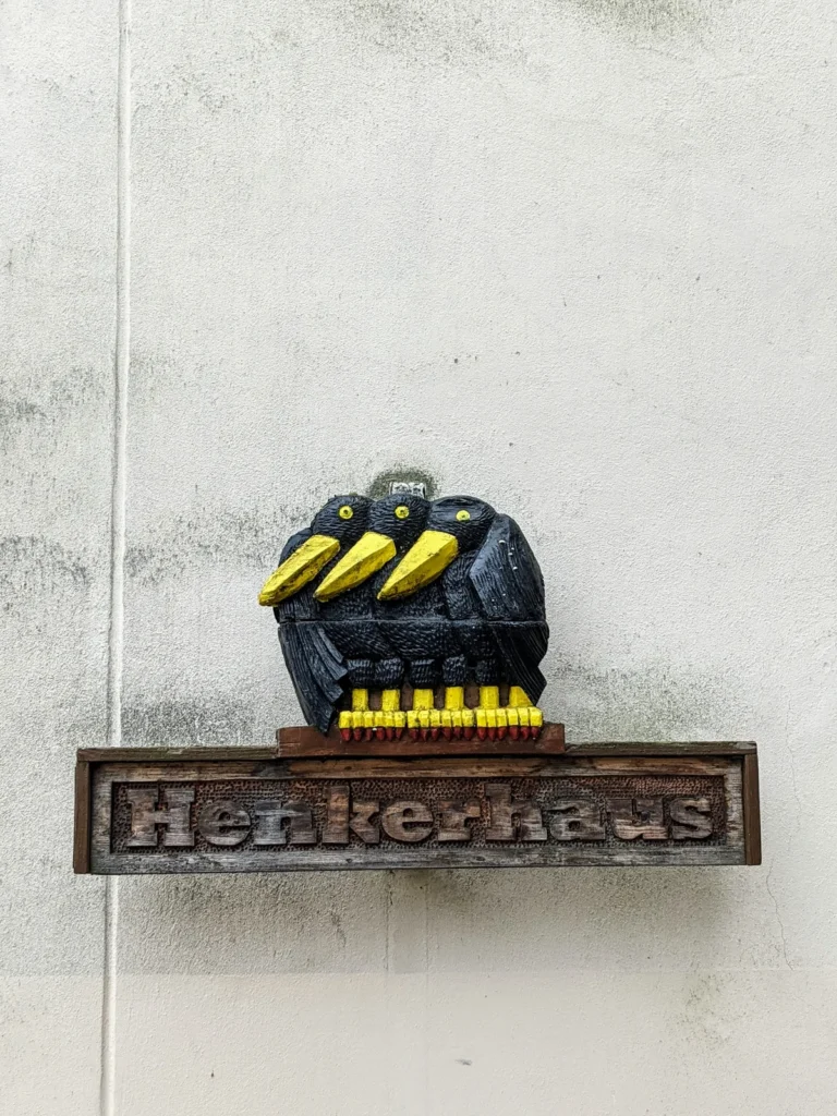 Schild „Henkerhaus“ mit drei schwarzen Vögeln mit gelben Schnäbeln an einer weißen Wand in Bernau