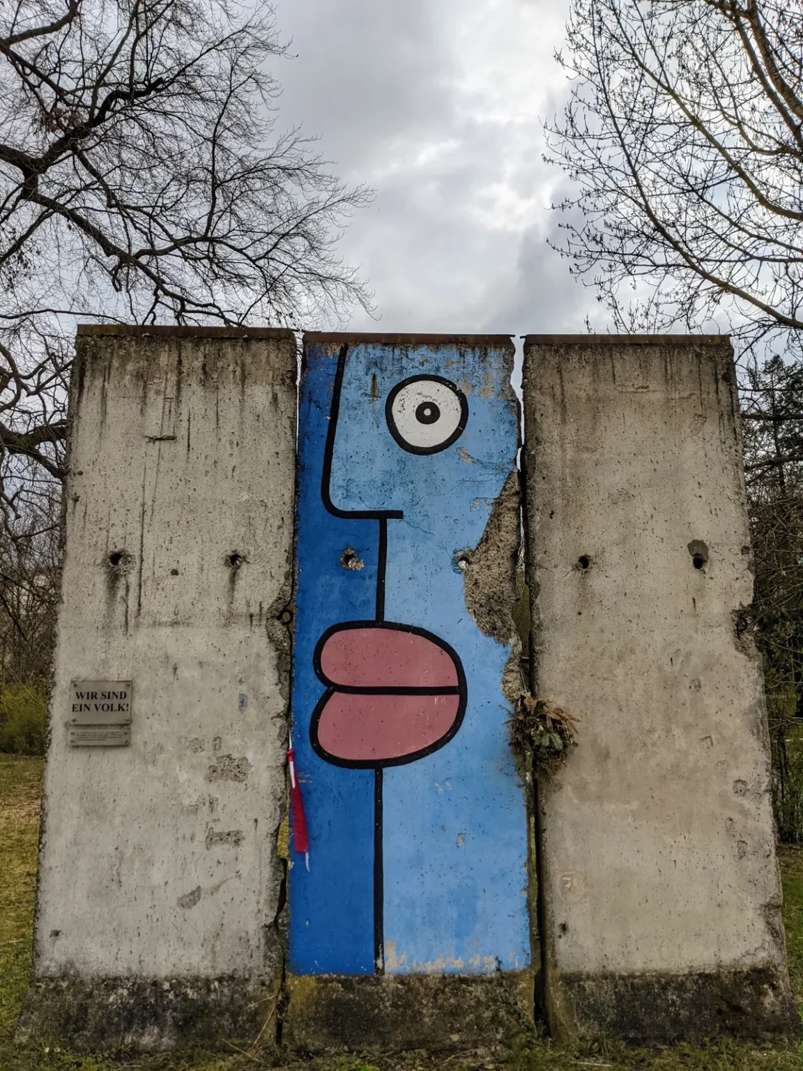 Von Thierry Noir bemaltes blaues Mauersegment mit Gesicht zwischen zwei unbemalten Segmenten in Bernau