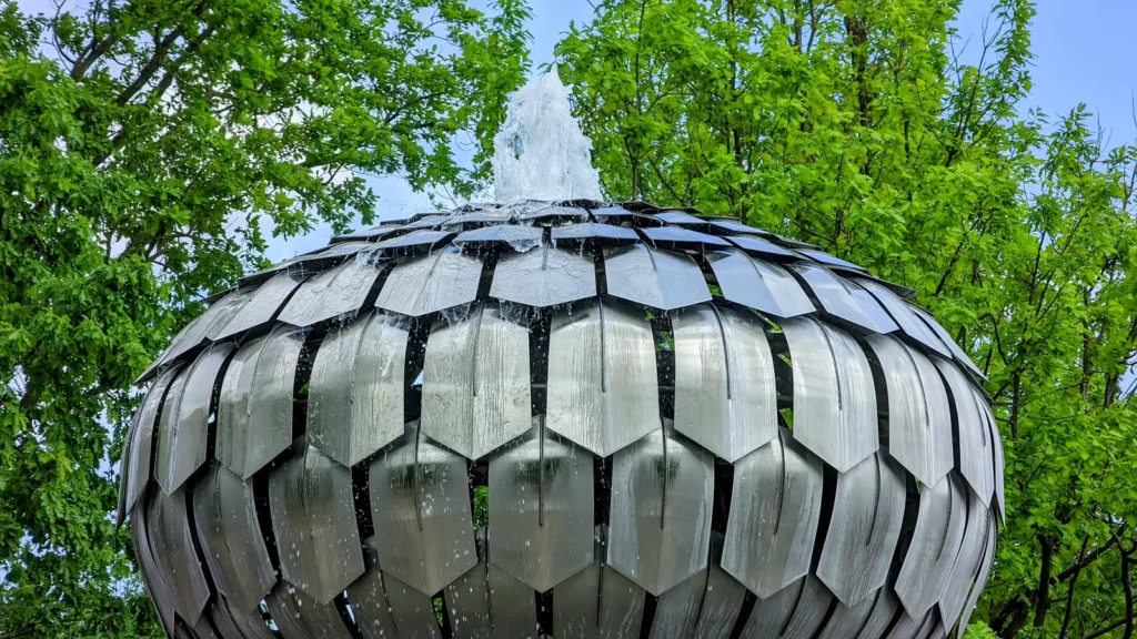 Kugelbrunnen aus Metallplatten mit Wasserfontäne vor Laubbäumen