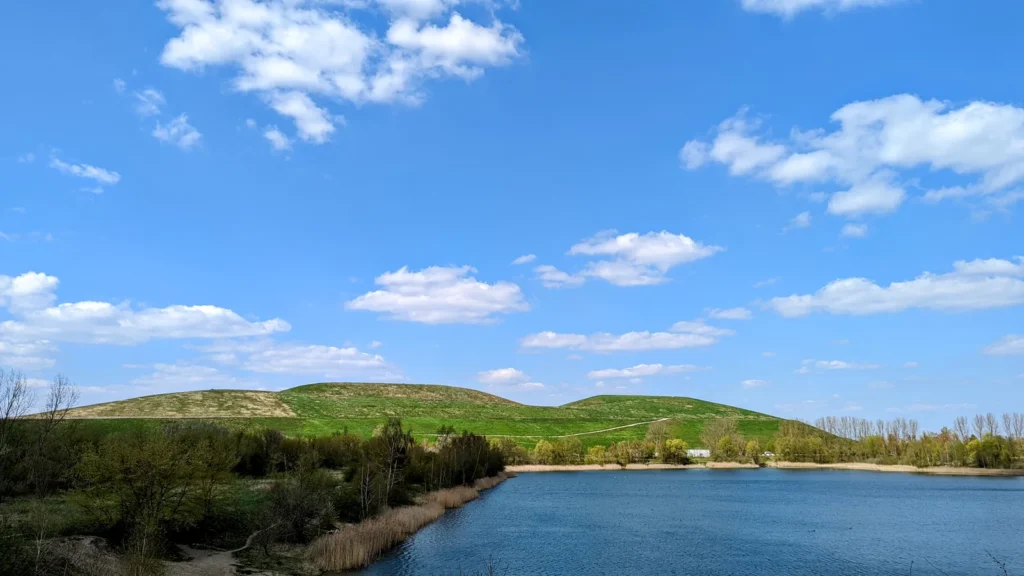 Blauer See vor zwei grünen Hügeln der Arkenberge in Blankenfelde, am Ufer Schilf und junge Bäume