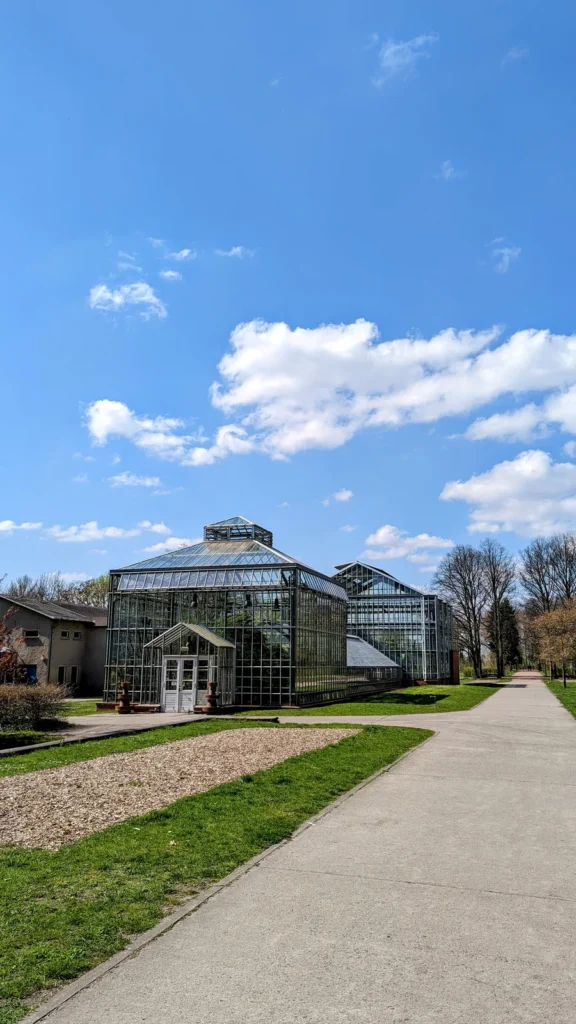 Historische Gewächshäuser mit Glasfassade im Botanischen Volkspark Blankenfelde, davor ein Betonweg und Rasenflächen