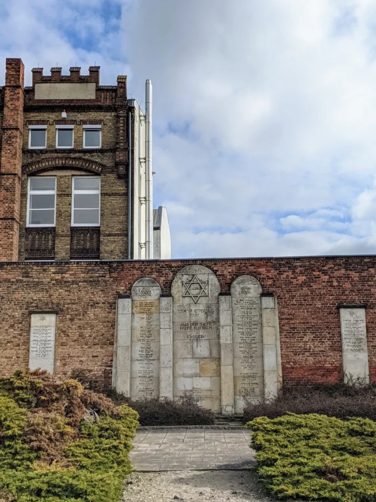 Gedenkwand aus Grabsteinen auf dem alten jüdischen Friedhof in Brandenburg an der Havel, dahinter ein Fabrikgebäude aus Backstein