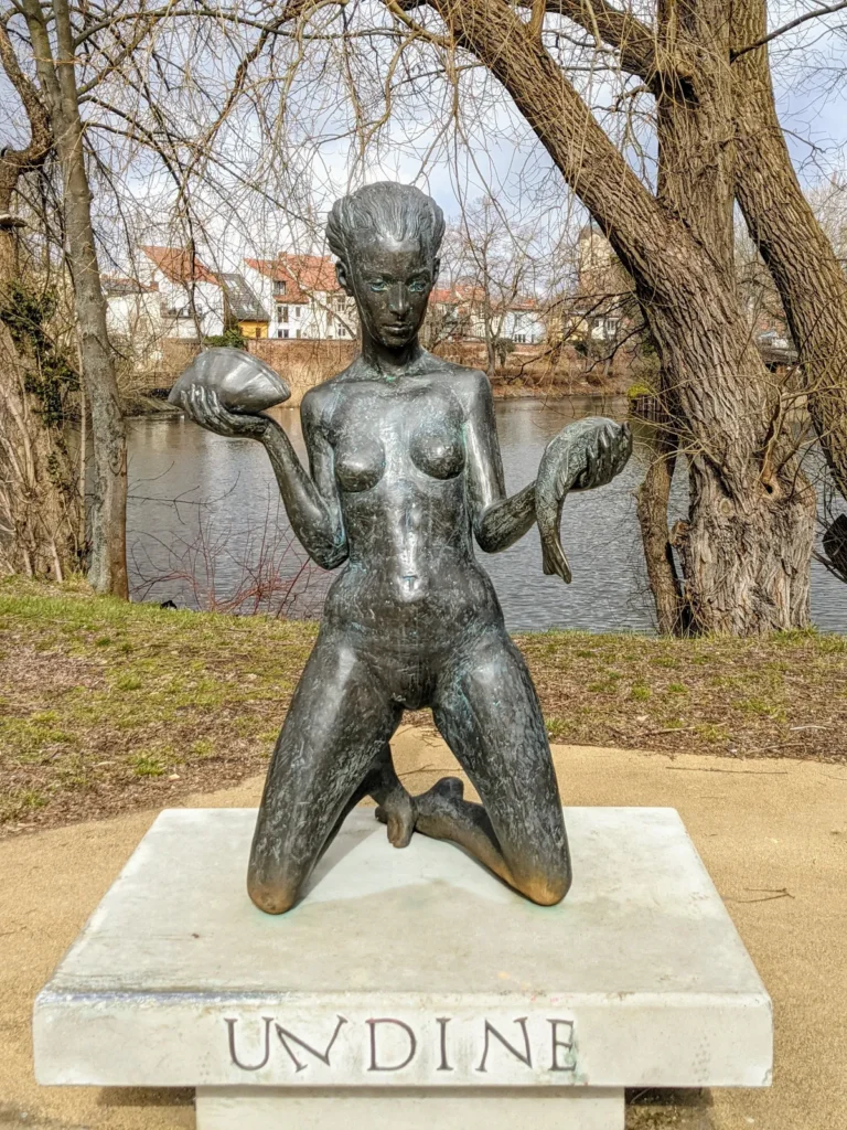 Bronzefigur Undine am Packhof, eine kniende Frau mit Fisch und Muschel in den Händen, im Hintergrund die Havel