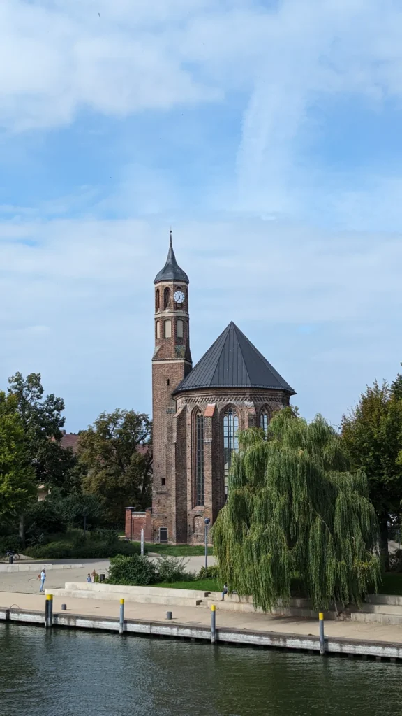 Kirche St. Johannis am Salzhofufer mit Backsteinchor und schlankem Turm, davor die Havel und eine Trauerweide