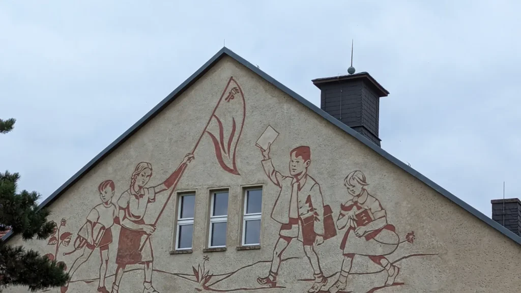 Giebel der Georg-Klingenberg-Schule in Brandenburg an der Havel mit rotbraunem Sgraffito: vier Schulkinder, ein Mädchen schwenkt einen Pionierwimpel
