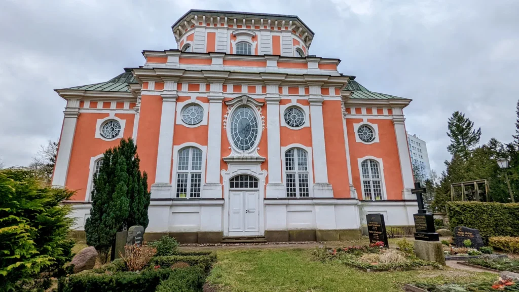 Frontansicht der Schlosskirche Buch mit orangeroter Fassade und Rundfenstern, rechts der Friedhof