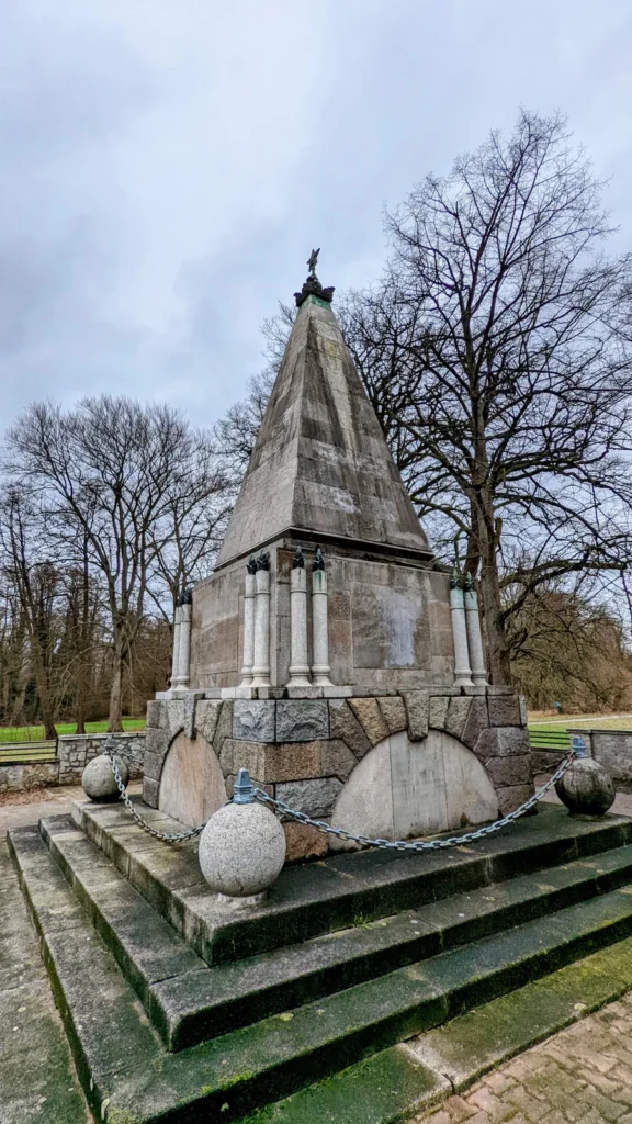 Pyramidenförmiges Ehrenmal auf Steinsockel mit Säulen und Kugeln an Ketten im Schlosspark Buch