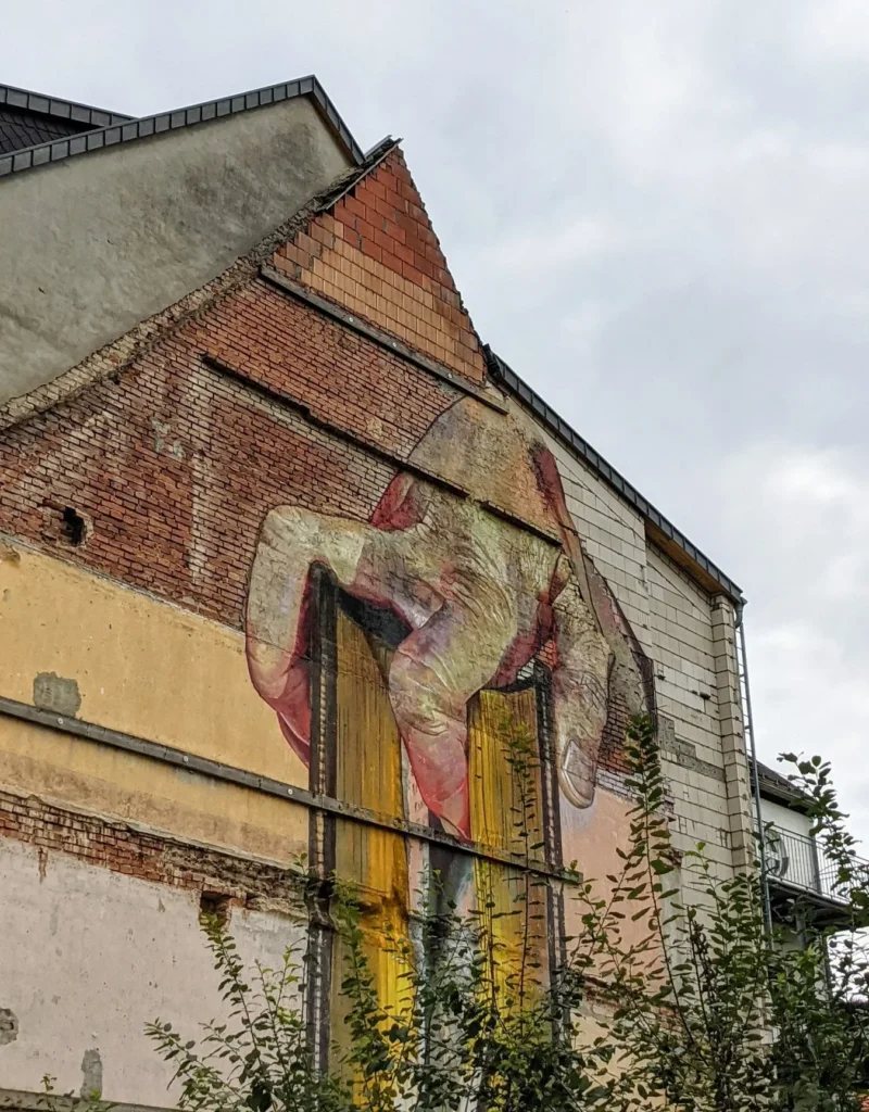 Wandbild von Case Maclaim an der Collegienstraße in Lutherstadt Wittenberg: gebeugte Figur in zarten Rosatönen auf einer Wand aus Backstein und Putz