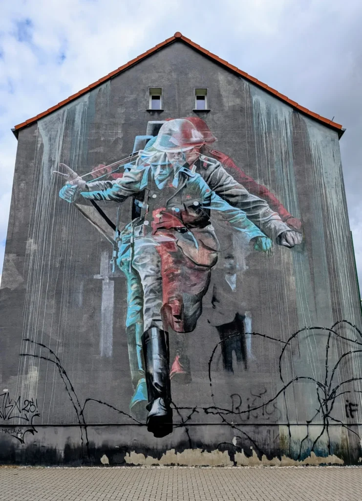 Wandbild Sprung in die Freiheit von Case Maclaim an der Sternstraße in Lutherstadt Wittenberg: schwebende Figur in zerrissener Kleidung über Stacheldraht auf grauer Giebelwand