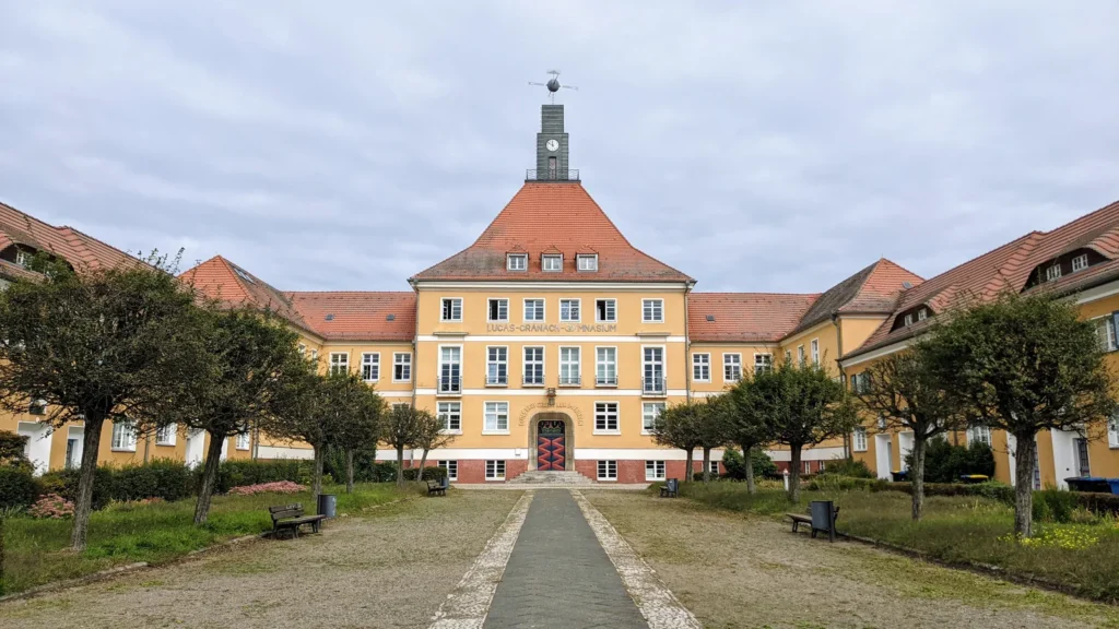 Ehemaliges Rathaus der Werkssiedlung Piesteritz, gelber Bau mit Turm und Seitenflügeln hinter einem Vorplatz mit Bäumen