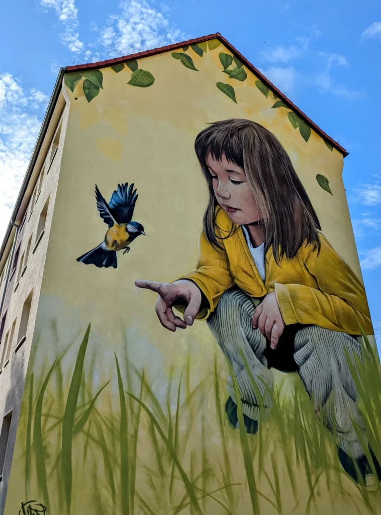 Wandbild Die Unschuld von Demut an der Odinstraße in Berlin-Karlshorst: Mädchen in gelber Jacke hockt im hohen Gras und streckt die Hand zu einer Blaumeise aus