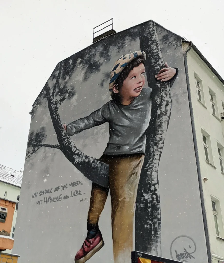 Wandbild von Demut an der Damerowstraße in Berlin-Pankow: Junge mit Mütze klettert an einem Baumstamm, daneben ein handgeschriebener Satz über Hoffnung und Liebe