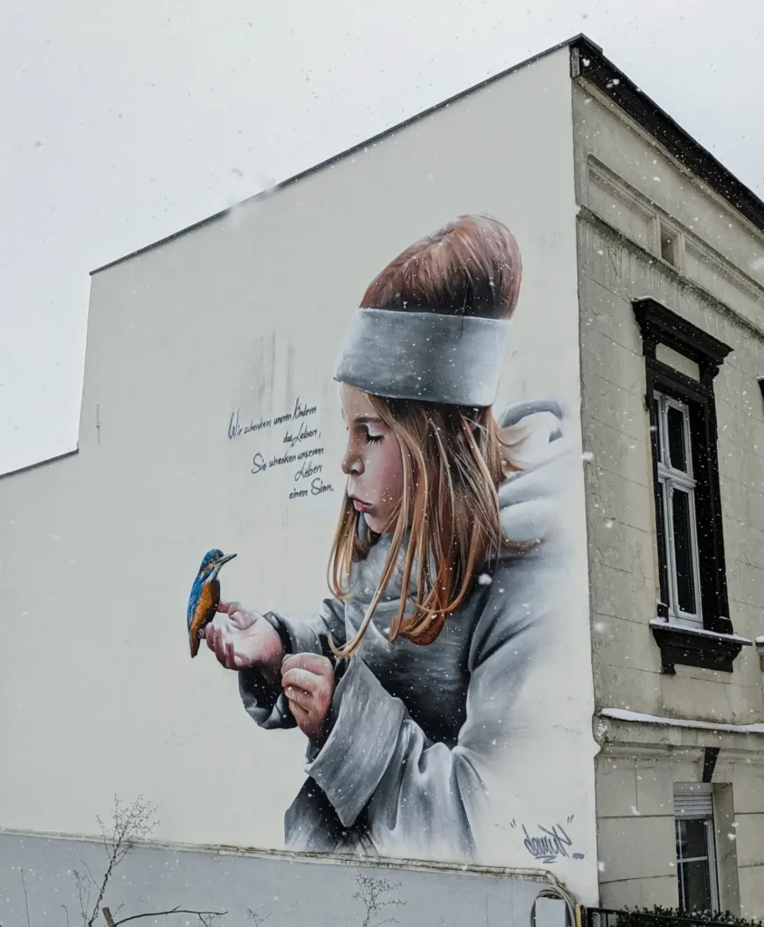 Wandbild von Demut an der Damerowstraße in Berlin-Pankow: Mädchen mit weißem Stirnband hält einen Eisvogel auf der Hand, daneben ein Schriftzug
