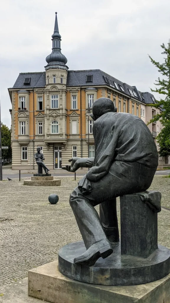 Bronzefiguren von Brecht und Weill auf dem Lidiceplatz in Dessau, dazwischen eine Kugel, im Hintergrund ein Eckhaus mit Turm
