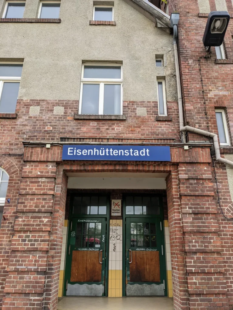 Eingang des Bahnhofs Eisenhüttenstadt, ein Klinkerbau mit blauem Stationsschild über grünen Türen