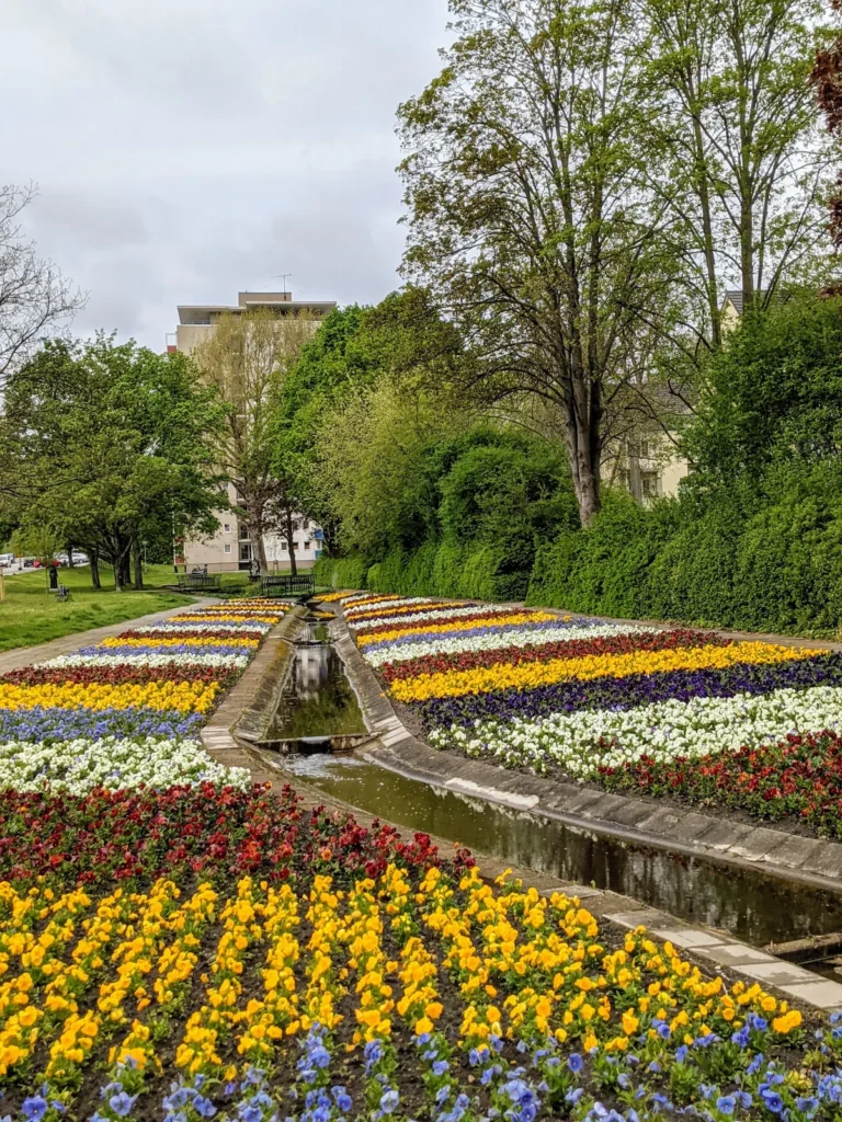Bunt bepflanzte Blumenbeete beiderseits einer schmalen Wasserrinne im Gartenfließ, dahinter Wohnhäuser zwischen Bäumen