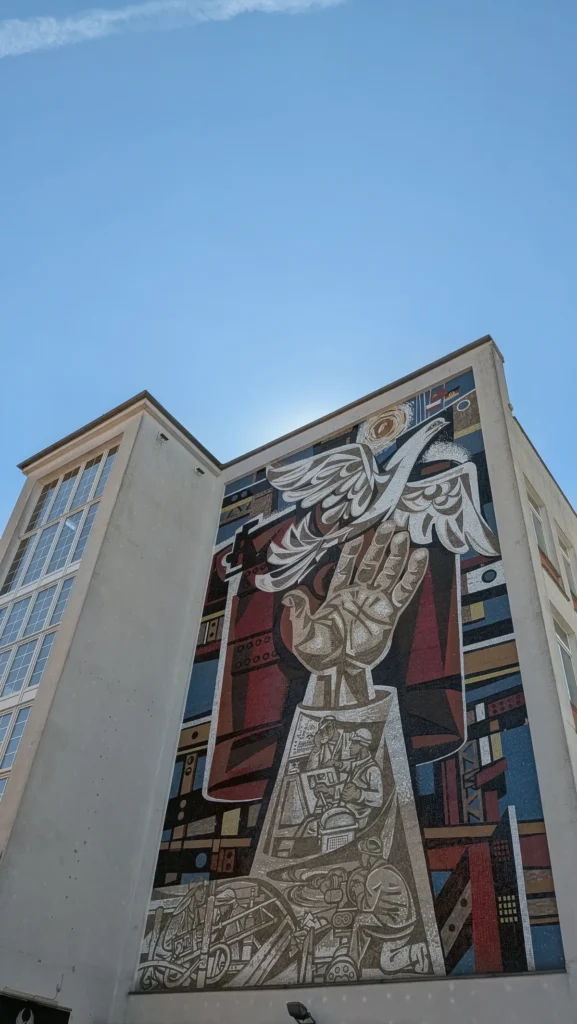 Wandmosaik von Walter Womacka am Lindenzentrum: eine große Hand hält eine weiße Taube, darunter Arbeiter und Industrie in Rot und Blau