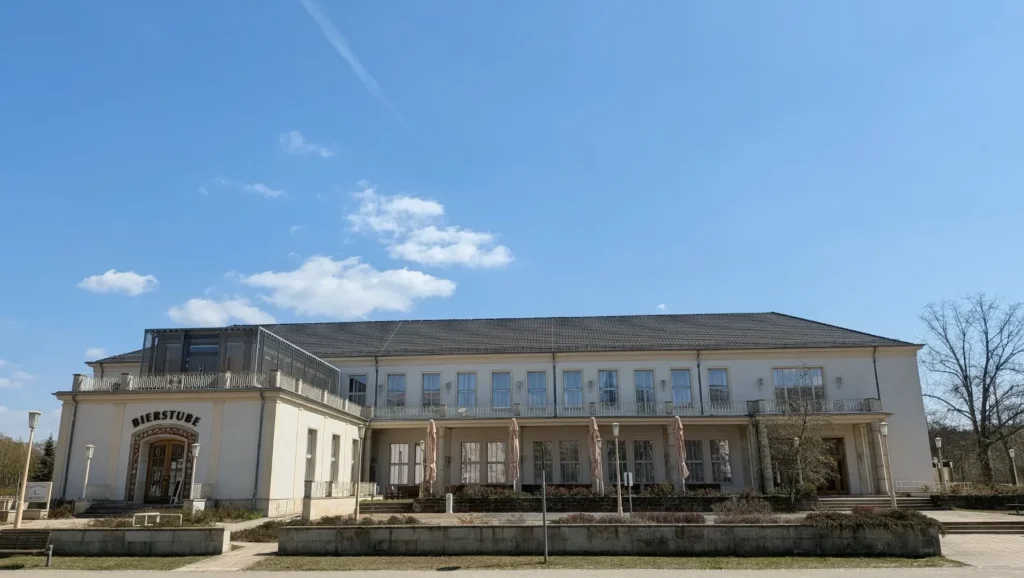 Zweigeschossiger heller Bau mit Terrasse, Pfeilern und einem Rundbogeneingang mit der Aufschrift Bierstube
