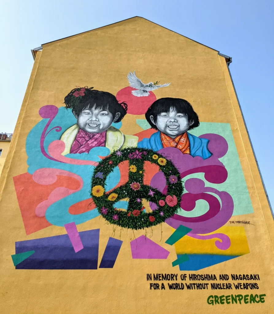Wandbild von Eme Freethinker für Greenpeace an der Köpenicker Straße in Berlin-Kreuzberg: zwei lachende Kinder, Friedenstaube und Peace-Zeichen aus Blumen auf gelber Giebelwand