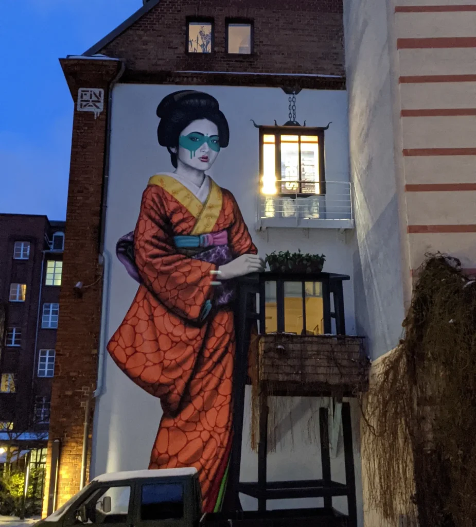 Wandbild Engeika von Fin DAC an der Köpenicker Straße in Berlin-Kreuzberg: Figur im orangen Kimono mit türkiser Maske an einer Hoffassade mit Balkon und Laterne