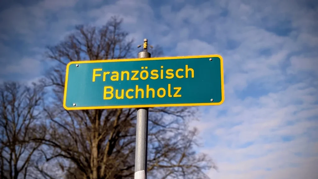 Türkisgrünes Ortsschild Französisch Buchholz an einem Mast vor kahlen Bäumen und Wolkenhimmel
