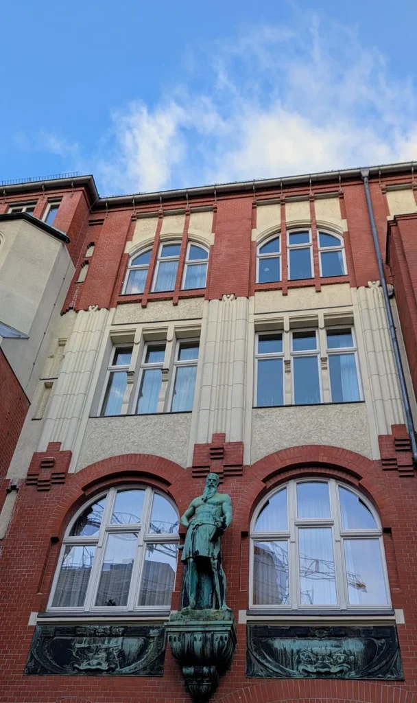Backsteinfassade mit Rundbogenfenstern und einer Bronzefigur eines bärtigen Mannes über einer Konsole in Friedenau