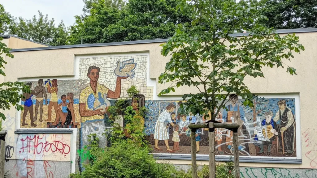 Flacher Bau mit Wandbild von arbeitenden Menschen hinter Bäumen im Sewanviertel in Friedrichsfelde