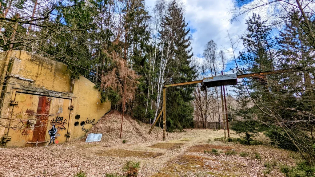 Gelber Betonbau mit rotem Tor auf einer Waldlichtung, rechts ein Portalkran