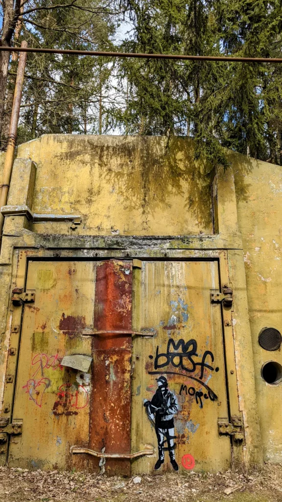 Gelbes Stahltor mit Rostbalken an einem Betonbau, daneben Graffiti und eine schwarze Figur