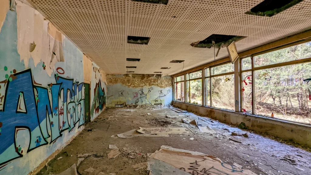 Raum mit blauem Graffiti an der Wand, Lochdecke und Fensterfront zum Wald