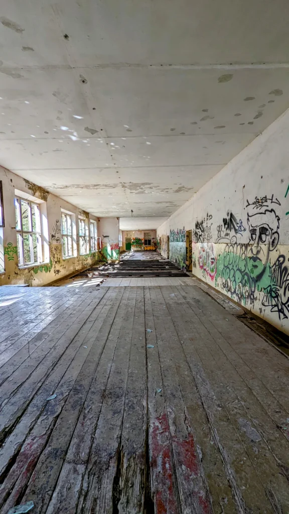 Langer Raum mit Dielenboden, Fensterreihe links und Graffiti an der rechten Wand, Hochformat
