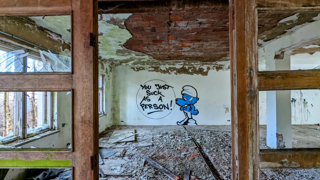Blick durch einen Holzrahmen auf das Graffiti einer blauen Zeichentrickfigur mit Sprechblase „You just suck as a person“