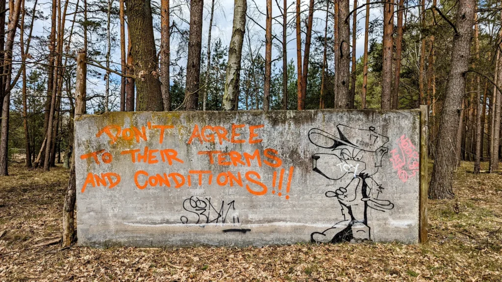 Betonwand im Kiefernwald mit orangem Schriftzug „Don’t agree to their terms and conditions“ und schwarzer Tierfigur