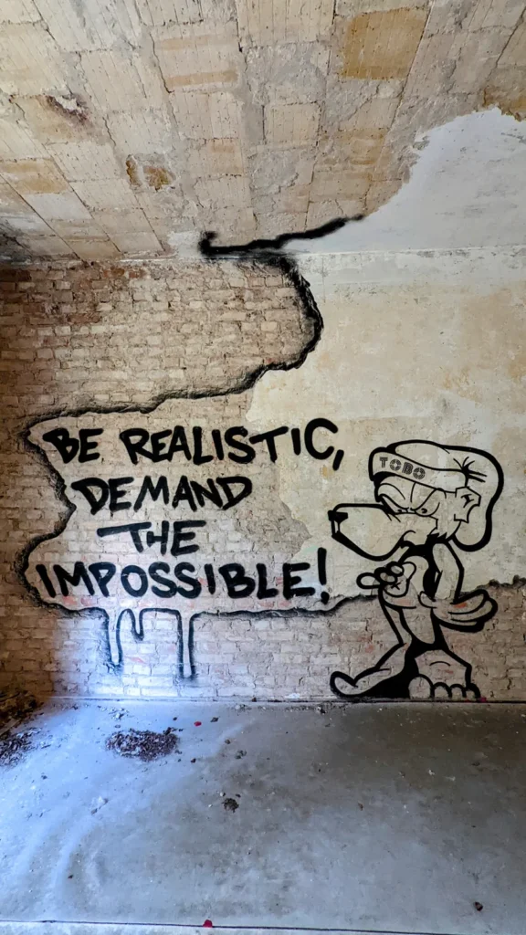Backsteinwand mit Sprechblase „Be realistic, demand the impossible!“ und Tierfigur