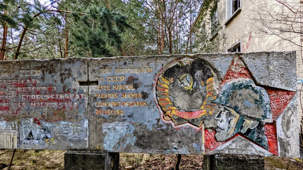 Betonrelief mit kyrillischer Schrift, Wappen und Soldatenköpfen zwischen Bäumen