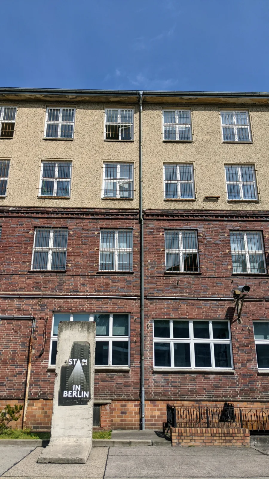 Backsteingebäude an der Gedenkstätte Hohenschönhausen mit Mauersegment und Schriftzug „STASI IN BERLIN“, vergitterte Fenster