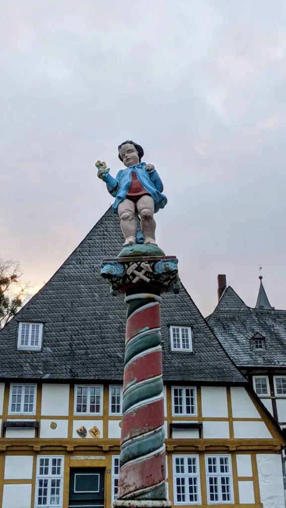 Bunt bemalte Figur auf einer rot-grün gestreiften Säule mit Bergbauzeichen vor Fachwerkhäusern