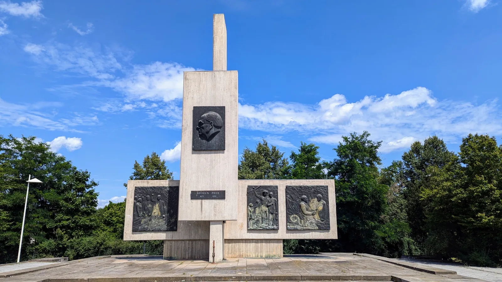 Wilhelm-Pieck-Denkmal in Guben: hohe Betonstele mit Bronzeporträt, davor ein Betonblock mit drei Bronzereliefs
