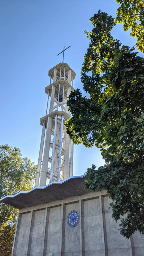 Offener weißer Glockenturm der Kaiser-Friedrich-Gedächtniskirche über dem geschwungenen Dach, daneben Laubbäume