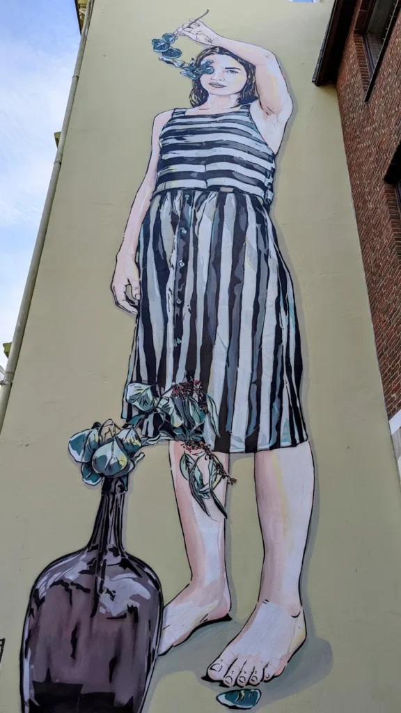 Wandbild von Jana und JS in der Amalienstraße in Hamburg-Harburg: Mädchen im gestreiften Kleid hält einen Eukalyptuszweig über den Kopf