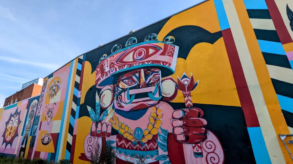 Wandbild von Ju Mu Monster an der Harburger Schloßstraße 36: bunte Maskenfigur mit großem Auge und Streifen in Gelb, Rosa und Blau