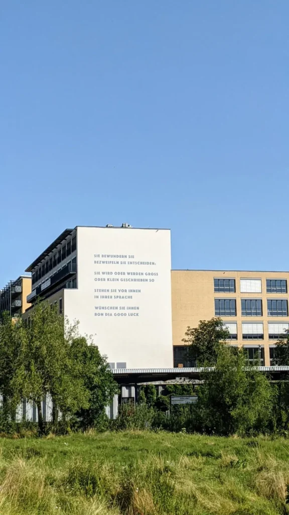 Weiße Giebelwand der Alice Salomon Hochschule in Hellersdorf mit Gedicht, davor eine Wiese