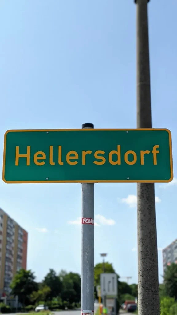 Grünes Ortsschild Hellersdorf an einem Mast, dahinter eine Straße mit Wohnhäusern