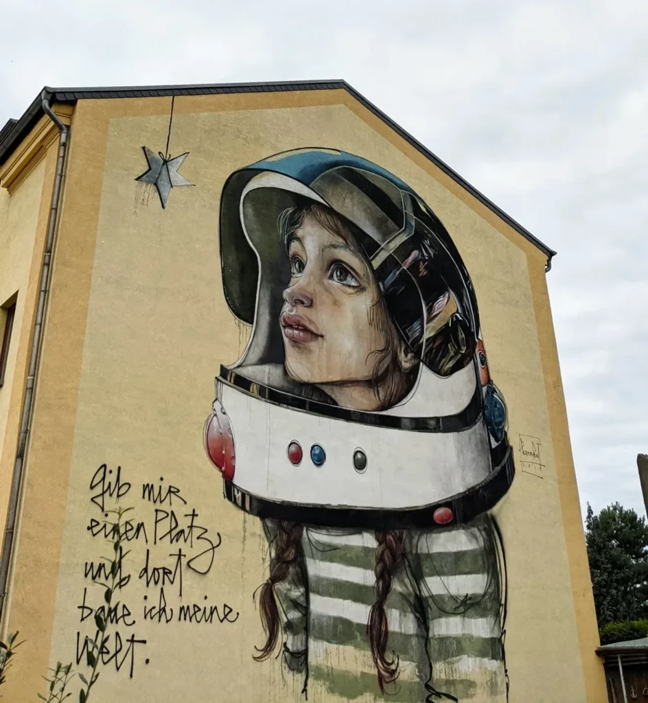 Wandbild von Herakut am Alten Bahnhof in Lutherstadt Wittenberg: Mädchen im Astronautenhelm neben einem Schriftzug an gelber Giebelwand