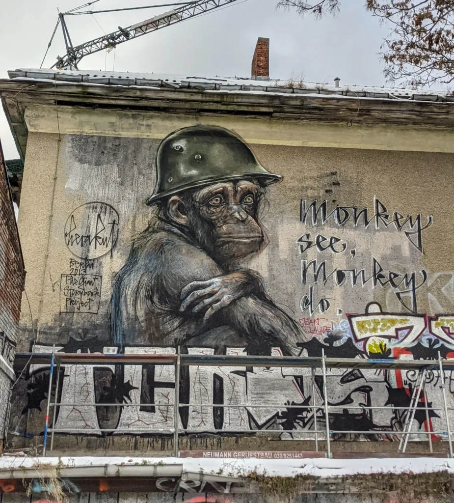 Wandbild Monkey see, monkey do von Herakut in Alt-Stralau in Berlin-Friedrichshain: Schimpanse mit Stahlhelm, daneben der handgeschriebene Titel