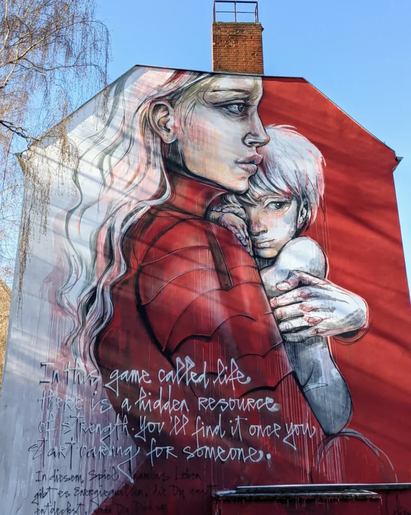 Wandbild This game called life von Herakut an der Sickingenstraße in Berlin-Moabit: Frau im roten Mantel mit gepanzert wirkenden Ärmeln umfasst ein Kind, daneben handgeschriebener Text