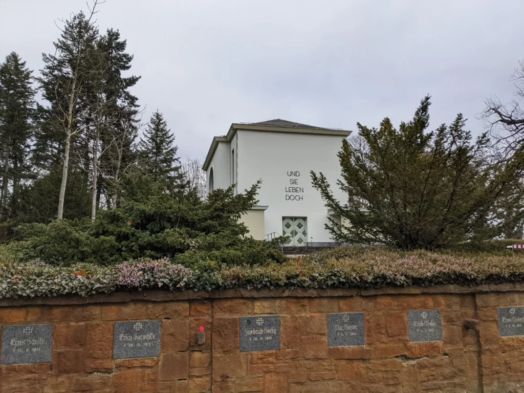 Feierhalle des Zentralfriedhofs Friedrichsfelde mit der Inschrift Und sie leben doch, davor die Sandsteinmauer mit Urnengräbern am Pergolenweg