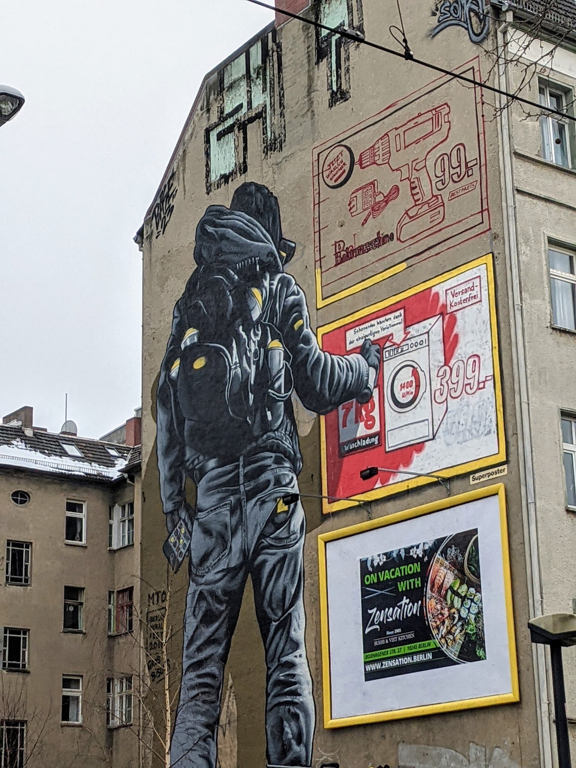 Wandbild von MTO an der Warschauer Straße: Ein Sprayer in schwarzer Kapuzenjacke sprüht vor gemalten Werbetafeln