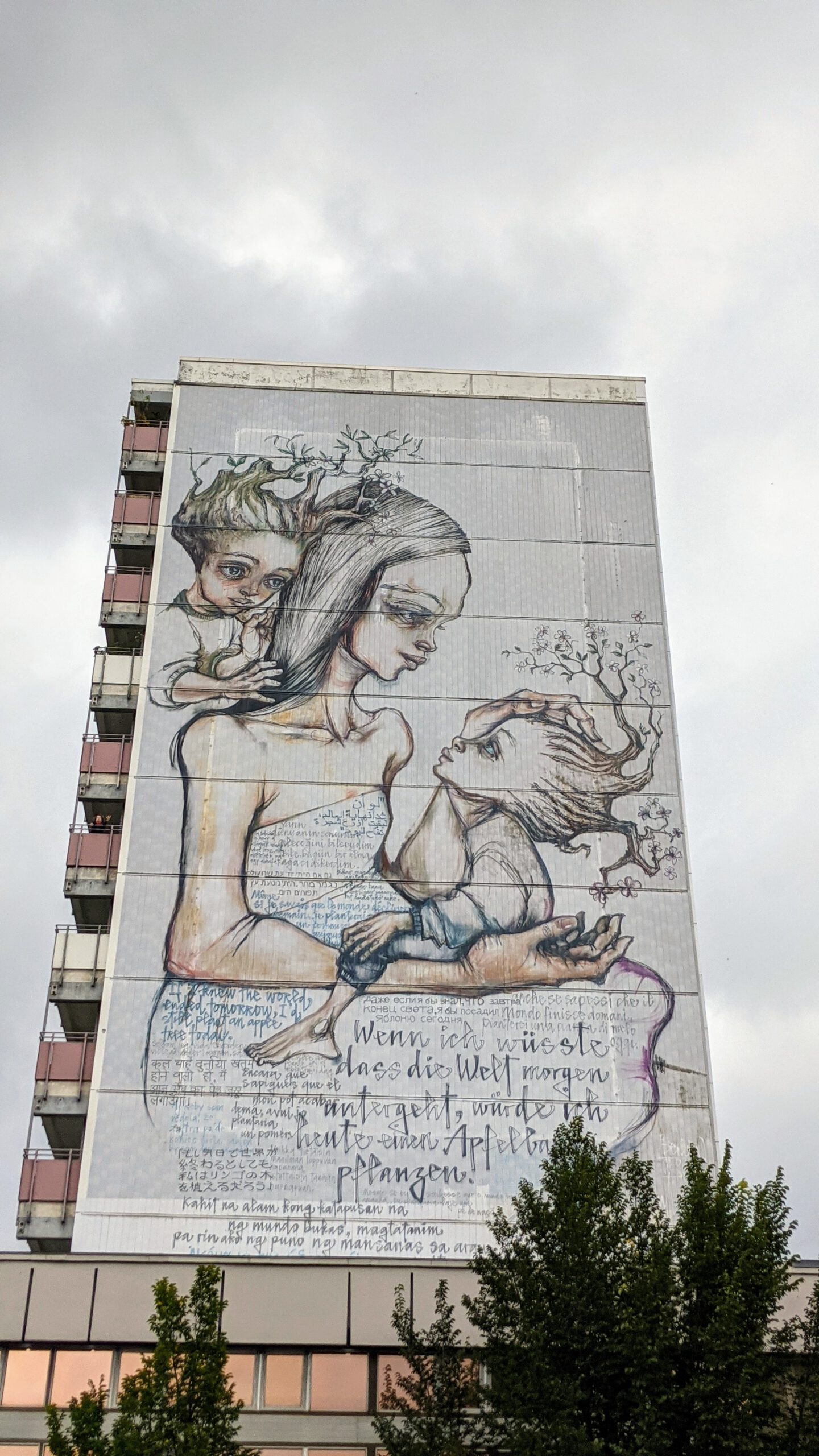 Riesiges Wandbild von Herakut an der Heinrich-Böll-Bibliothek: Eine Frau mit zwei Kindern, Zweige im Haar, darunter der Satz über den Apfelbaum in vielen Sprachen