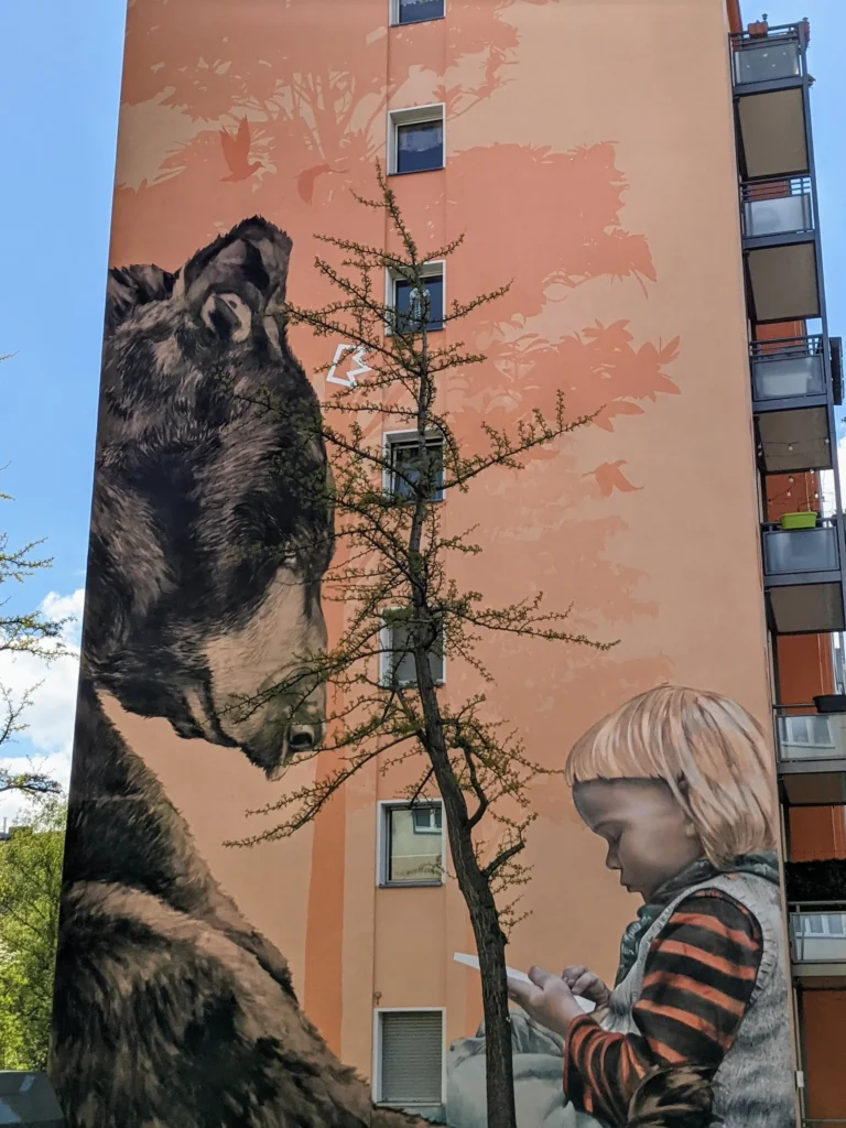 Wandbild Trialog von Innerfields an der Möckernstraße in Berlin-Kreuzberg: großer Bär neben einem Kind mit Tablet vor orangefarbener Fassade